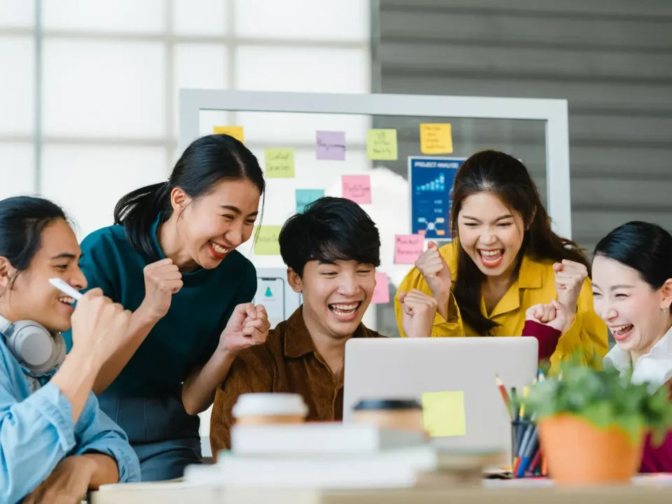 Tim Digital Agency yang kompak merayakan keberhasilan kampanye di depan laptop, menunjukkan efisiensi dan sinergi tim agensi dibanding in-house.